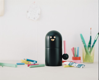Black toy figure with smiling face by JR–Work–Shop, surrounded by colorful pens and pencils