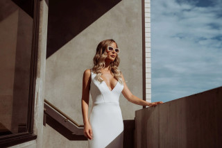 Woman in sunglasses and white dress stands on a concrete balcony